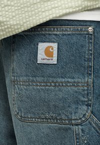 Πίσω τσέπη μπλε τζιν με μπαλώμα λογότυπο Carhartt, φορεμένη με πράσινο υφασμάτινο πουκάμισο και λευκό εσώρουχο.