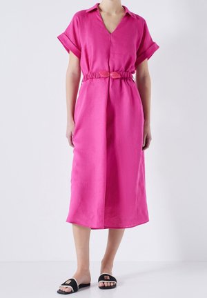 Lniana sukienka w kolorze fuksji z krótkimi podwiniętymi rękawami, dekoltem w serek i talią marszczoną, podkreśloną pasem w tym samym kolorze. Modelka zestawiła ją z czarnymi sandałami.