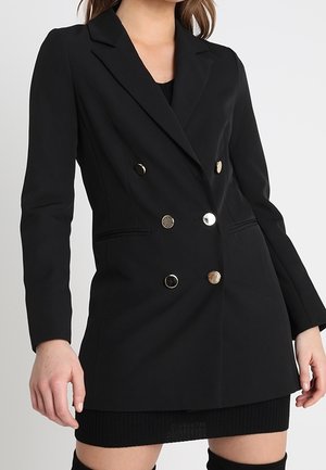 Kvinde iført en tætsiddende sort dobbeltbrystet blazer med seks metal knapper og et sort skørt mod en ensfarvet baggrund.