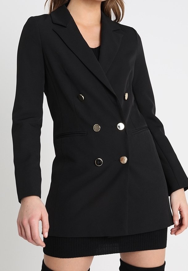 Femme portant un blazer noir cintré à double boutonnage avec six boutons métalliques et une jupe noire sur fond uni.