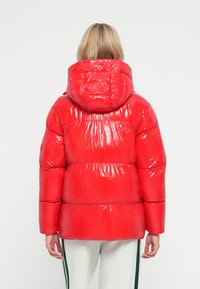 Veste matelassée rouge avec une finition brillante, dotée d'une grande capuche, d'une texture côtelée et de segments gonflés ; la vue arrière met en valeur la silhouette design.