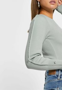 Long-sleeve, light green top featuring ribbed texture, subtle side seam detailing, and decorative button accents on the cuffs.