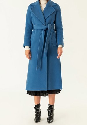 Femme portant un long manteau bleu ceinturé sur une jupe plissée noire et des bottines noires à lacets sur un fond clair.