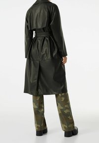 Persona vestida con un abrigo de cuero negro ceñido con cinturón sobre pantalones de camuflaje y zapatos de plataforma negros, de pie frente a un fondo blanco liso.
