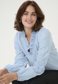 Light blue blouse with ruffled neckline and long, gathered sleeves. Textured fabric with subtle sheen and a keyhole opening at the front.