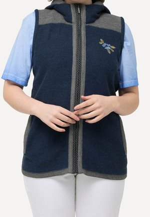 Person iført en marineblå og grå lynlåses uden ærmer vest med blomsterbroderi, over en lyseblå kortærmet skjorte og hvide bukser.