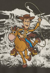 Gráfico de caricatura que presenta a un vaquero sobre un caballo marrón, con un lazo, usando un sombrero de vaquero, una camisa de cuadros y un pañuelo, sobre un fondo negro.