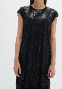 Černé šaty s krátkými rukávy, zdobené texturovaným vzorem z flitrů a detaily ve stylu třásní, což vytváří vrstvený efekt.