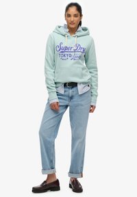 Jasnoniebieska bluza z kapturem z graficznym logo "Super Dry", zestawiona z podwiniętymi jasnoniebieskimi dżinsami i ciemnymi mokasynami. Widoczny kołnierzyk w paski.