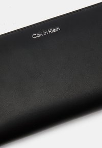 Mustassa nahkassa oleva lompakko, jossa on sileä pinta ja kohokuvioitu "Calvin Klein" -logo hopeana. Kompakti, tyylikäs muotoilu ilman näkyvää metalliosaa.