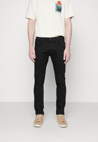 Schwarze Slim-Fit-Jeans aus Denim, mit Vorder- und Hintertaschen, genähten Nähten und einem klassischen Knopfverschluss. Kombiniert mit einem cremefarbenen T-Shirt.