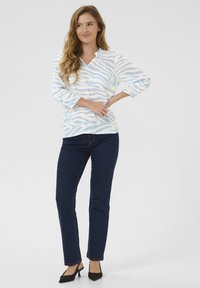 Blusa blanca con patrón de rayas azules, escote en V y mangas de tres cuartos, combinada con jeans ajustados azul oscuro y tacones negros.