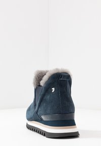 Botte cheville en suede bleu marine avec un revers en fausse fourrure, semelle rayée blanche et beige, et semelle extérieure en caoutchouc noir texturé. Présente un accent en logo argent.