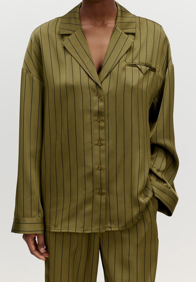 Personne portant un ensemble chemise en satin vert olive à manches longues et pantalon avec fines rayures verticales noires et un détail de poche pliée sur la poitrine.