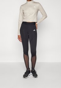 Beige sportovní top s dlouhým rukávem a jemným vzorem, kombinovaný s černými legínami se síťovanými panely a bočními pruhy. Černé běžecké boty dokládají celý outfit.