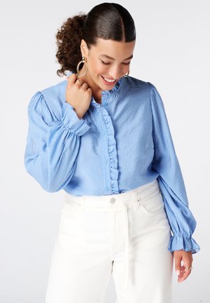 Vrouw die glimlacht en naar beneden kijkt, draagt een blauw gestreepte blouse met ruches, witte broek en grote gouden oorringen met krullend haar naar achteren gebonden.