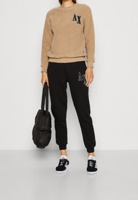 Felpa in pile beige con logo, pantaloni della tuta neri con logo sulla gamba, borsa nera e sneakers nere con dettagli bianchi.