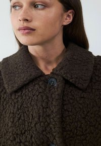 Bruine gestructureerde jas van een dikke, fuzzy stof, met een grote kraag en twee zwarte knopen aan de voorkant. Close-up van de schouder en de neklijn.