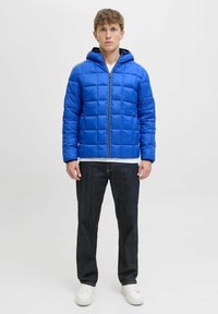 Junger Mann steht mit nach vorne gerichtetem Blick und trägt eine blaue gesteppte Kapuzenjacke, dunkle Jeans, weiße Turnschuhe und ein weißes Hemd unter der Jacke.