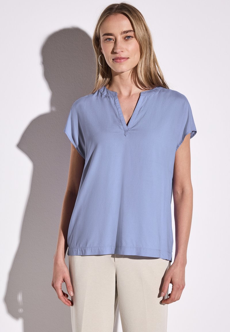 Blusa blu chiaro a maniche corte con scollatura a V, in tessuto morbido, vestibilità comoda e orlo dritto. Il modello indossa pantaloni color beige.
