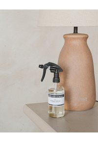 Flacone spray trasparente etichettato "Voyage Vétiver" accanto a una base lampada in terracotta alta e curvata su una superficie e parete beige.