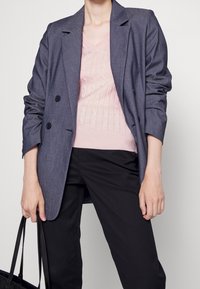 Blazer grigio doppiopetto con revers a punta, indossato sopra un maglione a trecce rosa chiaro, abbinato a pantaloni neri e una borsa a tracolla nera.