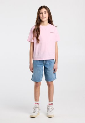 Meisje met lang bruin haar, een roze shirt, blauwe denim short tot op de knieën, witte sokken met rode strepen en witte sneakers.