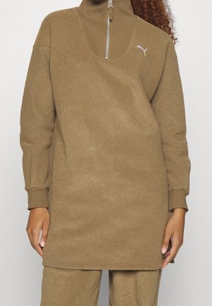 Kobieta ubrana w brązowy pulower z długim rękawem, z przednim zamkiem błyskawicznym i logo Puma na piersi, w połączeniu z pasującymi brązowymi spodniami.