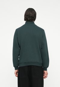 Donkergroen sweatshirt met een hoge kraag, ribbelmanchetten en een relaxte fit, weergegeven vanuit de achterkant tegen een effen witte achtergrond.