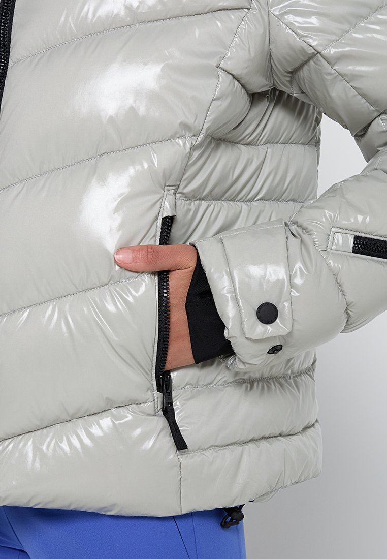 Hellgraue glänzende gesteppte Jacke mit schwarzem Reißverschlussfach, Hand im Taschenfach und am unteren Ende sichtbare blaue Hose.