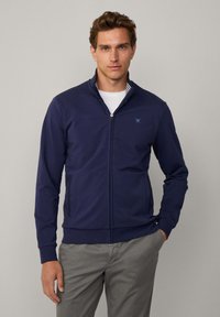 Giacca a zip blu navy realizzata in un materiale morbido, con colletto alto a righe all'interno e due tasche laterali.