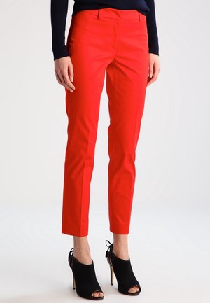 Personne portant un pantalon slim court rouge vif et des chaussures à talons hauts noires ouvertes à l'avant, debout devant un fond uni.