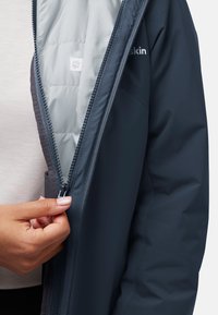 Une veste bleu marine possède une doublure en tissu matelassé gris. La fermeture éclair est partiellement ouverte, mettant en valeur une texture douce et une étiquette avec un motif de patte à l'intérieur.
