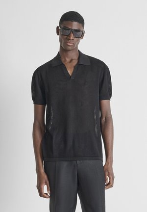 Man wearing black mesh short-sleeve polo shirt, black pants, and large black sunglasses, standing against plain light background.