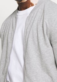 Uomo che indossa una T-shirt bianca a girocollo sotto una giacca leggera grigia con zip, che mostra una condizione della pelle sul collo e sul basso viso.