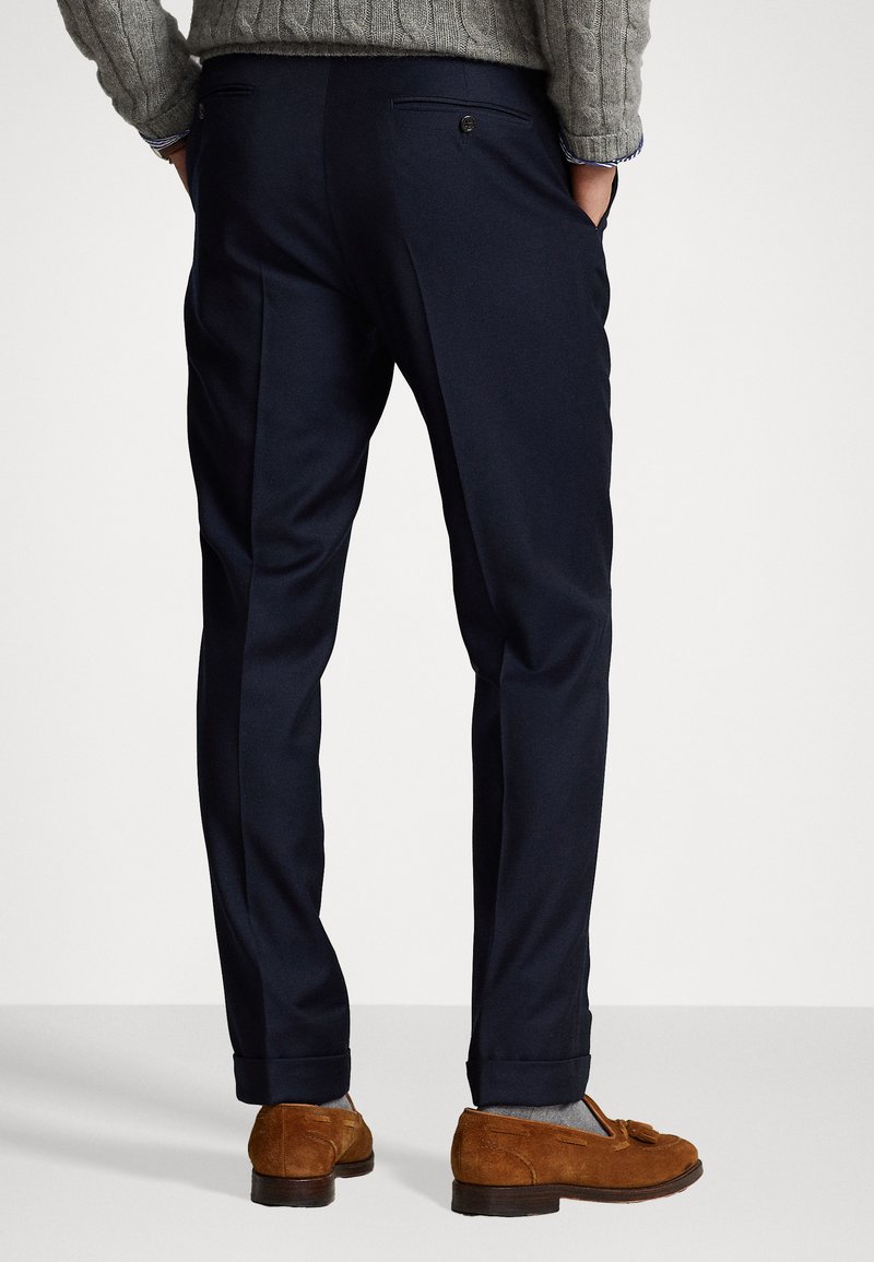 Pantalons ajustés bleu marine en tissu lisse, dotés de poches arrière avec boutons et de revers. Assortis à des mocassins en daim marron.