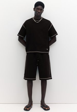 Tânăr stând pe fundal alb, purtând un tricou negru oversized și pantaloni scurți cu margine albă, papuci negri și coliere cu mărgele.