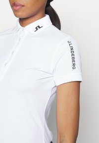 Hvit polo skjorte med korte ermer, med en svart logo på kragen og "J.LINDEBERG" tekst langs skuldersømmen. Myk stofftekstur.
