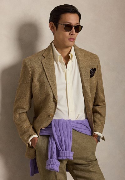 Mees, kes kannab pruuni tekstuuriga pintsakut, valget nööbitavat särki, lilla kampsunit vöökohale seotud, tumedaid päikeseprille ja kella, käed taskutes.