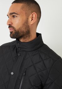 Veste matelassée noire avec col montant, fermetures à bouton et poche zippée sur la poitrine. Tissu texturé avec un motif en losanges.