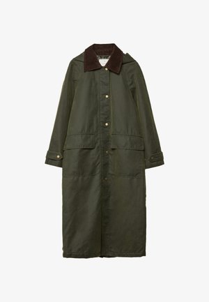 Person iført en mørkegrøn lang frakke, sort sweater, hvid lagdelt nederdel med blonderkant og sorte knæhøje støvler, stående mod en grå væg.