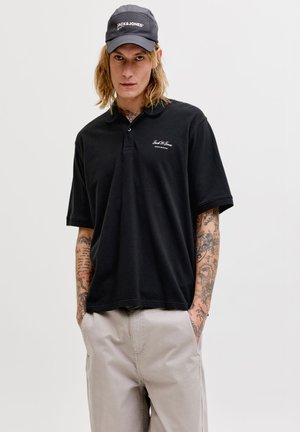 Άνδρας που φοράει μαύρο καπέλο Jack & Jones, μαύρο πόλο με λογότυπο και μπεζ παντελόνι, στέκεται με τα χέρια στις τσέπες μπροστά σε απλό φόντο.