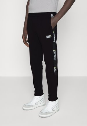 Persona con pantalones deportivos negros EA7 Emporio Armani con franjas de logotipo a los lados y zapatillas blancas de caña alta, de pie contra un fondo blanco.