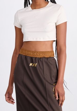 Kremfarvet crop top med korte ærmer og lille hvid Nike-logo. Brune sweatpants med elastik i taljen og gyldne "NIKE"-bogstaver.