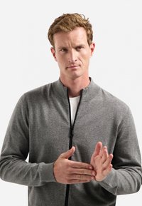 Man met lichtbruin haar die in een grijze ritssweater zijn handen klapt tegen een effen witte achtergrond.