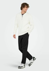Weißer Strickpullover mit Reißverschlusskragen, kombiniert mit schwarzen Hosen und schwarz-weißen Sneakers, Modell läuft vor einem schlichten Hintergrund.