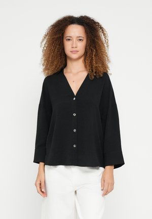 Blouse noire à boutons avec un col en V, manches trois-quarts et tissu texturé. Comprend quatre boutons ronds sur le devant.