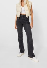 Light beige puffer vest, sleeveless design, zip closure. Black high-waisted jeans, straight-leg fit. White sneakers with minimal detailing.