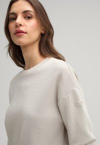Light beige sweatshirt with a crew neckline, soft texture, and subtle embroidered logo on the sleeve. Simple, relaxed design.