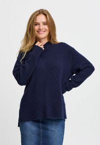 Dunkelblaues gestricktes Sweatshirt mit hohem Ausschnitt, lockerer Passform und langen Ärmeln. Strukturiertes Material und seitliche Schlitze am Saum. Kombiniert mit einem Jeansrock.